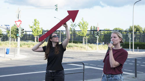 Zwei junge Frauen stehen an einer großen Kreuzung mit Ampeln. Fahrzeuge sind nicht zu sehen. Eine der beiden hält einen großen roten Pfeil schräg aufwärts gerichtet über dem Kopf. Die andere spricht in ein Mikrofon.