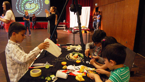 In einem großen Veranstaltungssaal sitzen vier Kinder an einem Tisch und basteln. Im Hintergrund sind Erwachsene zu sehen, die in die Hände klatschen.